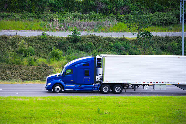Profile of rapid road beast in the form of modern powerful truck with a tough periphery and refrigeration unit on the trailer with caps on the wheels with unrivaled aerodynamic shape on a green summer road gives the illusion of speed of movement of the time.